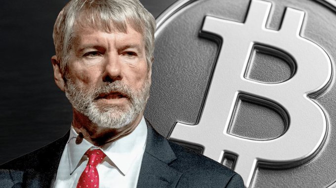 An older man with a white beard in a suit stands in front of a large silver Bitcoin symbol.