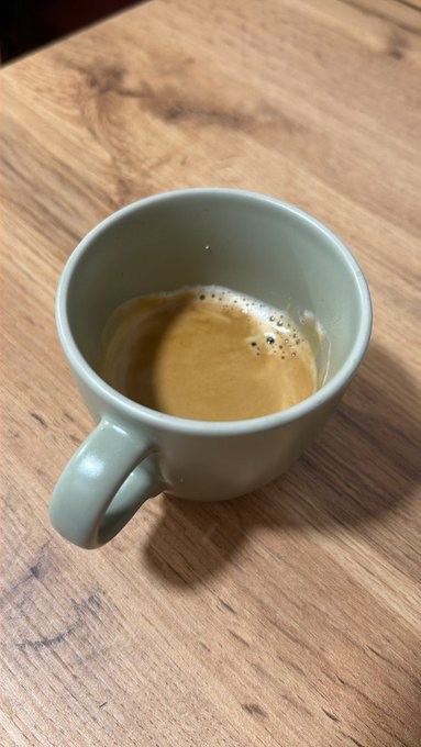 The image features a cup of coffee sitting on top of a wooden table. The cup has a light green color, which contrasts with the dark brown wood of the table. There are no other objects or people visible in the photo, making the focus solely on the cup and its surroundings.