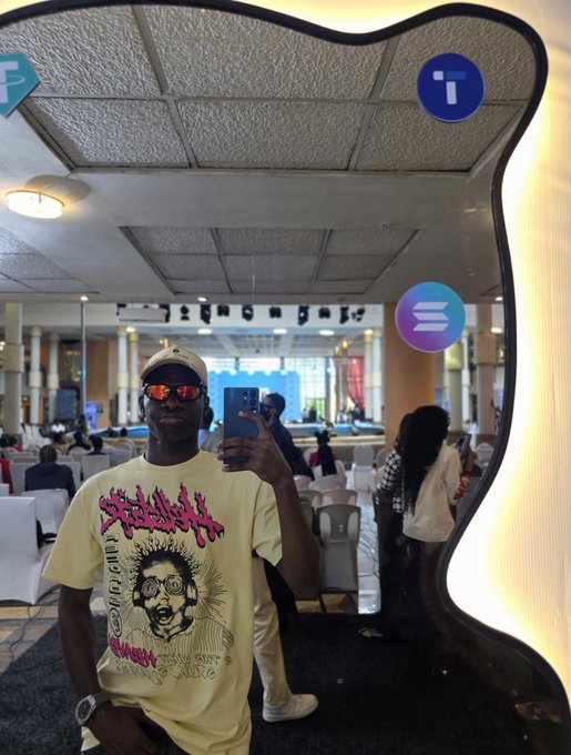 A mirror selfie of a young man wearing a beige graphic t-shirt, sunglasses, and a baseball cap, holding a phone in what appears to be a banquet hall or conference center.