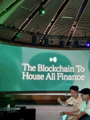A view of a presentation or panel discussion with a large screen displaying 'The Blockchain To House All Finance,' with audience members visible in the foreground.