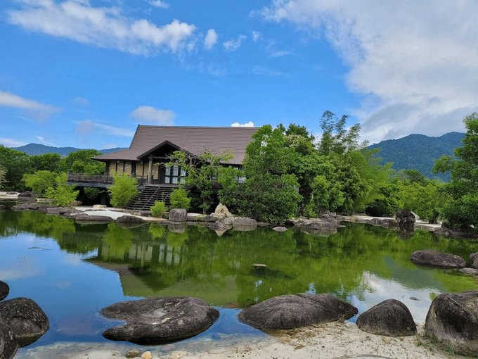 A serene photograph of a traditional Japanese-style house with a brown tiled roof, situated beside a calm pond reflecting green trees, with large stones arranged along the water's edge under a blue sky.
