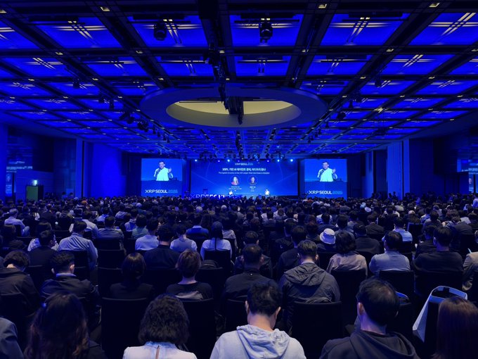 A large, dark conference hall is filled with an audience facing a brightly lit stage, with blue lighting overhead and "XP SQUARE" displayed on the screens.