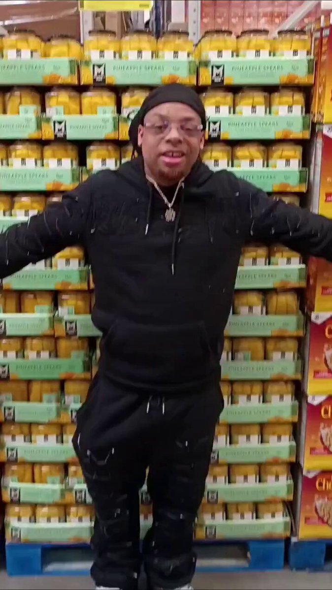 iced tea boxes rest on a shelf, with a man in black attire standing behind them and a yellow sign reading "A-Liquids" above him.