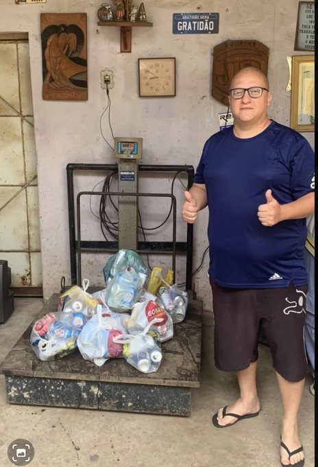 A man wearing glasses, a navy blue Adidas t-shirt, and brown shorts stands in a room giving two thumbs up next to several bags of recyclables on a scale. A sign reading 'QUE A TUDO SEJA DADO EM GRATIDÃO' is visible on the wall behind him.
