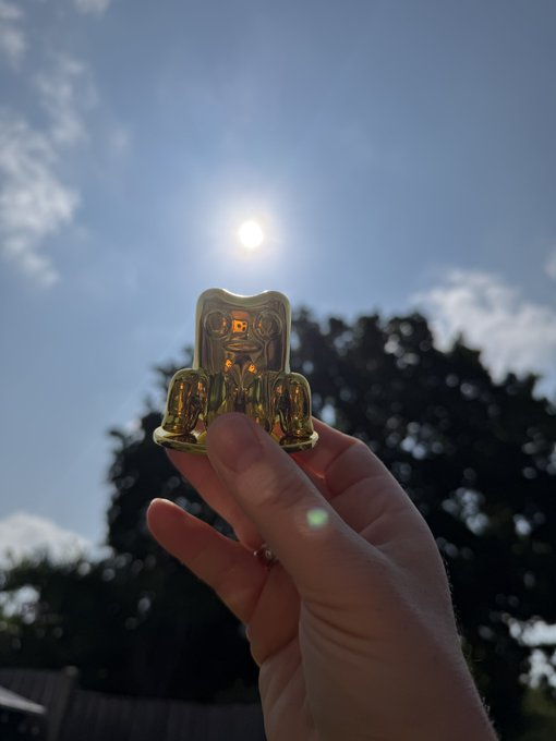 A hand holding up a small golden figurine against a bright blue sky with the sun shining behind it and trees in the background.