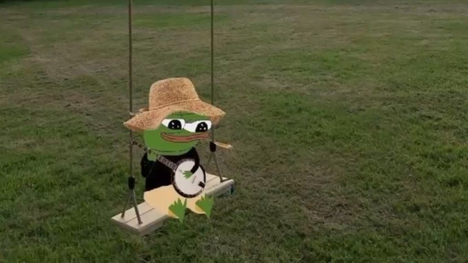 A green Pepe the Frog plush toy wearing a straw cowboy hat and holding a banjo, sitting on a wooden swing in a grassy field.