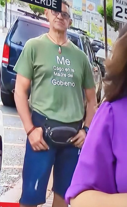 A man wearing sunglasses stands on a street wearing a green t-shirt that reads 'Me Cago en tu Madre del Gobierno' with a fanny pack around his waist.
