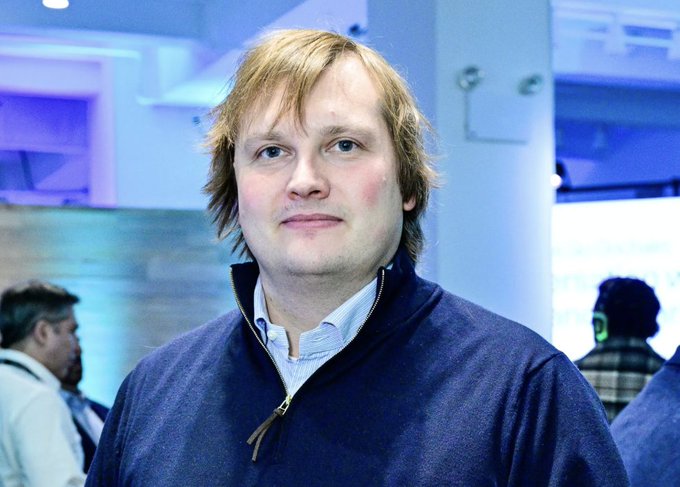 A man with tousled blonde hair, wearing a navy blue zip-up sweater over a light blue collared shirt, stands in what appears to be a brightly lit event space with blue-toned lighting. This image accompanies news coverage of Aave founder Stani Kulechov, who is flagging strong resistance to Ethereum's EIP-8361 proposal, suggesting it could rank among the most opposed changes in the network's history.
