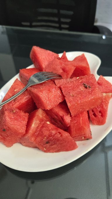 A white plate is piled high with cubed red watermelon, with a silver fork resting on top, placed on a dark glass table.
