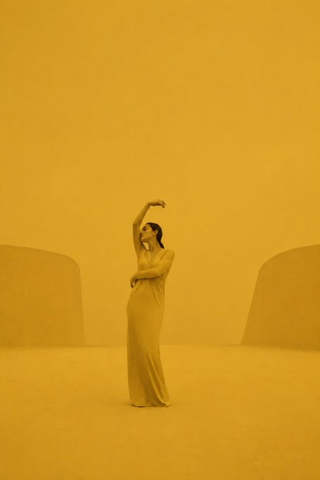 A woman in a flowing dress poses gracefully with one arm raised overhead in a minimalist yellow-toned studio space.