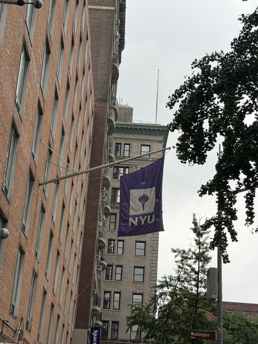 A purple NYU (New York University) banner hangs from a brick building, with a street sign for "Washington" visible below, near what appears to be Washington Square Park in New York City. The "Waddup @degens" caption suggests a casual greeting to fellow NYU students or locals, fitting the campus-area setting.
