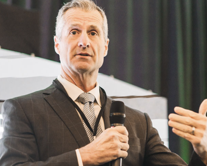 A middle-aged man with graying hair in a dark suit and tie speaking into a microphone at what appears to be a conference.