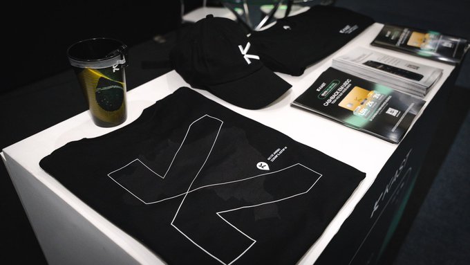 A photograph of Kai Cenat merchandise displayed on a white table, including a black t-shirt with a large outlined 'X' logo, a black cap with the same logo, a clear tumbler with a yellow design, and Kai Cenat branded packs.