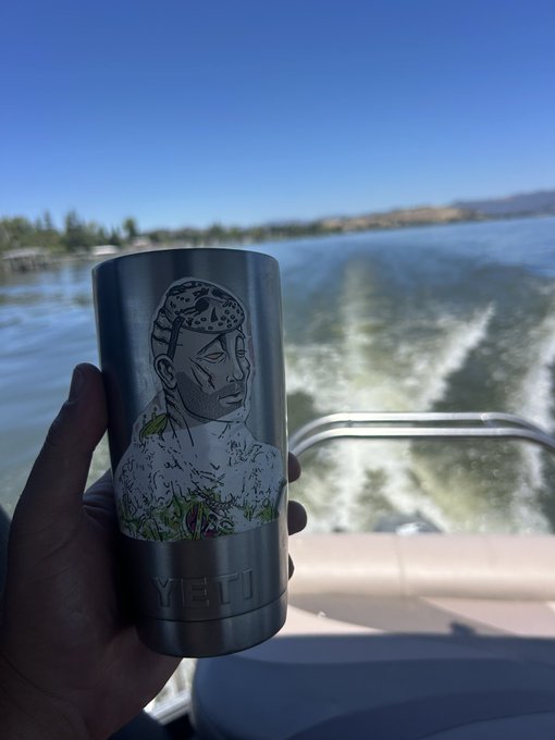 A hand holds a YETI tumbler featuring a colorful portrait design of a man with a patterned head covering, with the brand name "YETI" visible at the bottom, set against the backdrop of a boat's wake on a sunny lake. The scene captures a leisurely moment on the water, fitting the post's theme of long-time support for the DeGods NFT community.