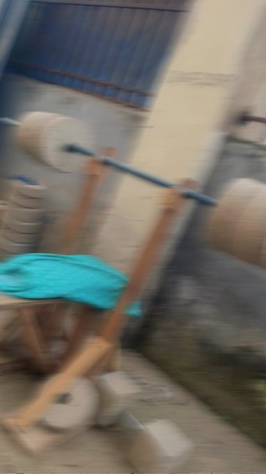 A blurry outdoor photograph showing what appears to be a makeshift workout setup with rolled materials, a wooden frame, and a teal cloth on a dirt ground near a building.
