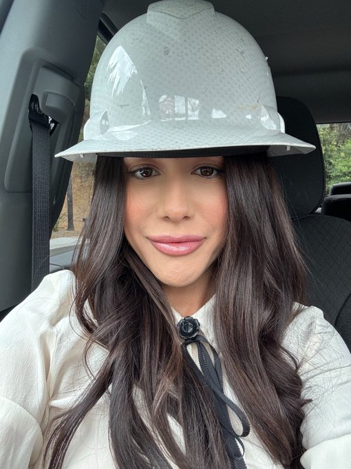 A woman wearing a white hard hat with "I ❤️ NY" stenciled on it sits in a car, dressed in a white top with a black neck tie. The image's caption playfully nods to the "savage" buying energy of altcoin season, suggesting she's suiting up like a worker ready to tackle the market head-on without setting limits.