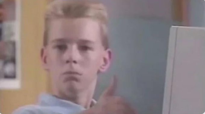 A young boy with a skeptical expression looking at a computer monitor, captured in what appears to be a vintage TV show or film screenshot.