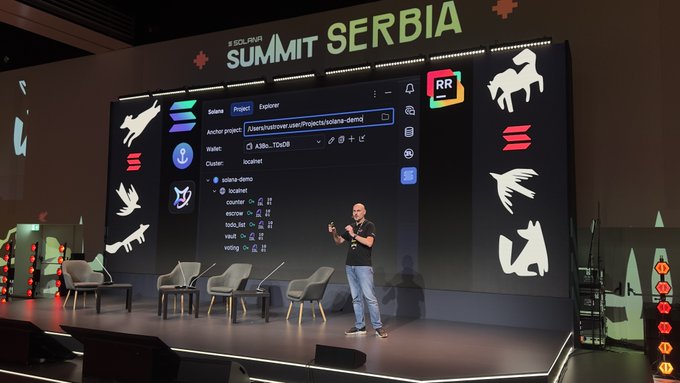 A presenter on stage at SOLANA SUMMIT SERBIA displaying a code interface (Solana Explorer) on a large screen behind him, with Solana-themed decorations.