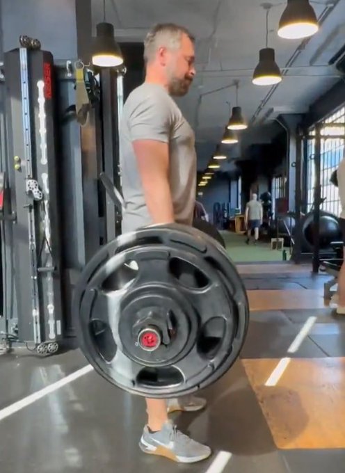 A bearded man in a gray t-shirt performs a heavy barbell deadlift in a gym, loading a large black weight plate onto the bar. The image humorously aligns with the caption "nobody: the Solana Founders:", visually exaggerating the founders' well-known physical fitness and bodybuilding lifestyle.