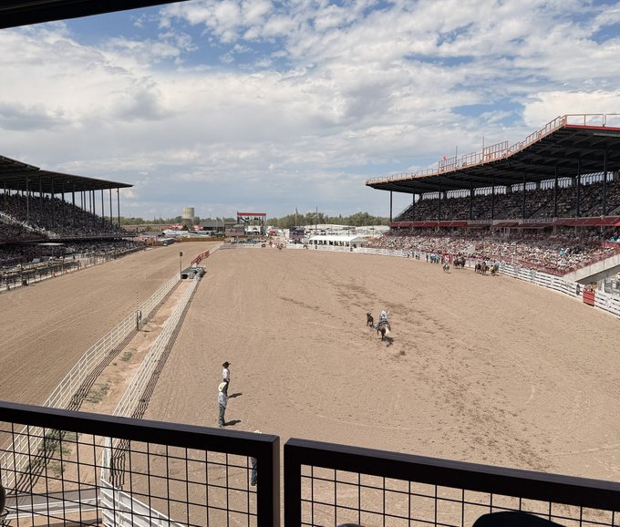 A wide view of a sunlit rodeo arena where two riders on horseback move across the dirt floor, with packed grandstands stretching along both sides under a partly cloudy sky—evoking the wide-open, high-stakes spirit behind "Long bitcoin and rodeos."