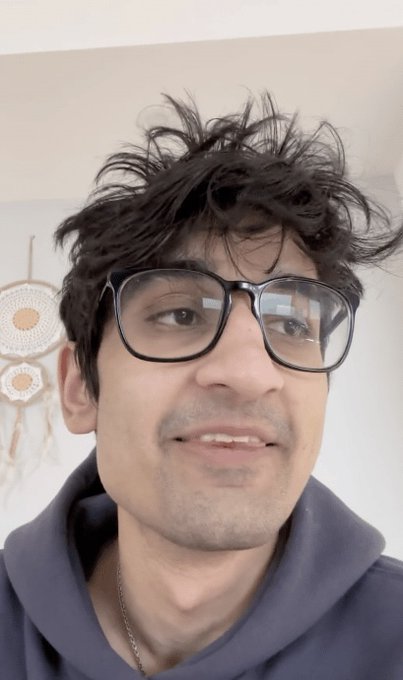 A close-up selfie of a young man with messy dark hair wearing black-rimmed glasses and a dark gray hoodie.