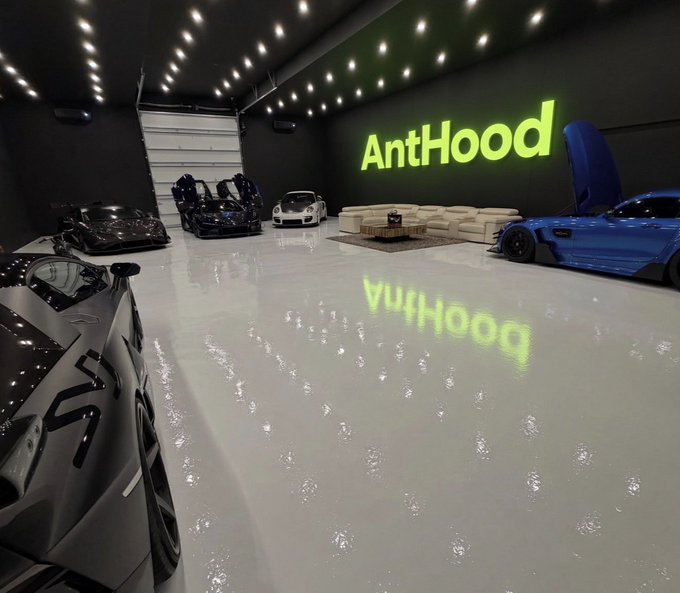 A luxurious garage showroom displays several high-end sports cars—including a blue Mercedes-AMG GT and a white Porsche—arranged around a seating area, with a prominent illuminated green "AntHood" sign on the back wall. The sleek black interior with glossy floors reflects the glowing logo, embodying the brand's bold, defiant message of permanence and luxury presence.