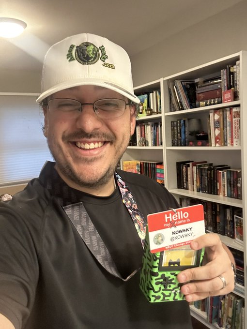 A man wearing glasses and a white cap smiles while holding a "Hello My Name Is" tag labeled "KOWSKY," standing in front of a bookshelf.