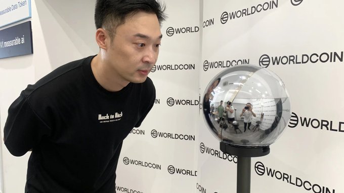 A man wearing a "Black in Black" t-shirt leans toward a mirrored orb at a Worldcoin booth, where the "WORLDCOIN" logo repeats across the backdrop. The image visually echoes the post's warning about people queuing for iris scans in exchange for Worldcoin (WLD) tokens, highlighting how the company's branded devices collect biometric data at events.