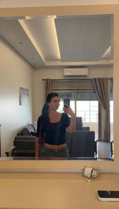 A young woman with long dark hair, wearing a black top and green pants, takes a mirror selfie in a modern living room with an air conditioner visible in the reflection, while her phone displays a "gm" (good morning) message—matching the caption's upbeat morning greeting to those who echo positivity back.