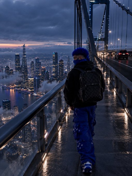 A person with blue hair wearing a black jacket, backpack, and blue patterned pants stands on a bridge overlooking a city skyline with illuminated skyscrapers and traffic at dusk.
