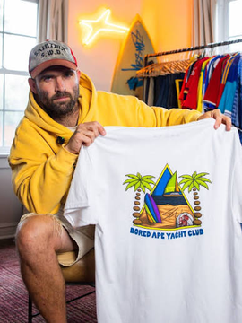 A man in a yellow hoodie and cap holds up a white t-shirt featuring the Bored Ape Yacht Club logo with palm trees and a surfboard design, promoting the brand's new Sean Wotherspoon collab. The neon "Ape" sign and clothing rack of colorful merch in the background reinforce the surf-culture drop the post references.