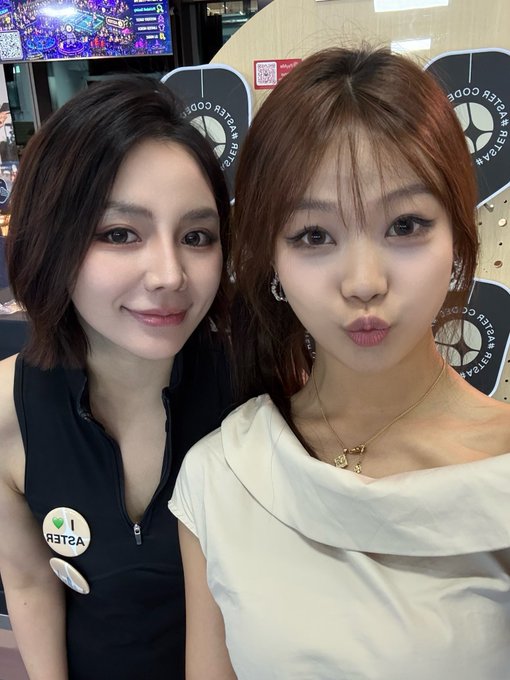 Two women pose for a selfie at what appears to be a K-pop venue, with one wearing a black vest with 'ASTERO' visible and the other in a white off-shoulder top, with 'PLANETARY CODE' signs in the background.