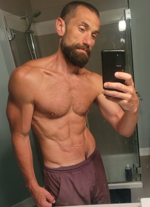 A bearded, muscular shirtless man takes a mirror selfie with his phone while wearing maroon shorts, displaying his defined abs and athletic physique in what appears to be a bathroom or gym setting. The image visually reinforces the post's message about Bitcoin enthusiasts embracing healthier, fitness-focused lifestyles.