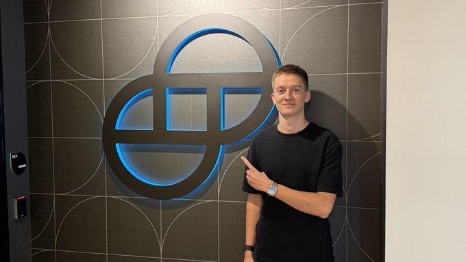 A young man in a black t-shirt points at a large illuminated blue logo on a dark wall.