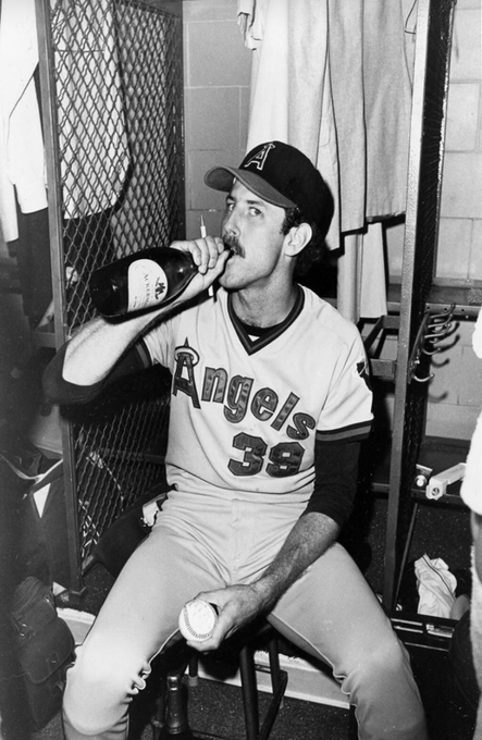 A black-and-white photo of a baseball player wearing an Angels jersey with the number 38, sitting in a locker room and drinking from a bottle.