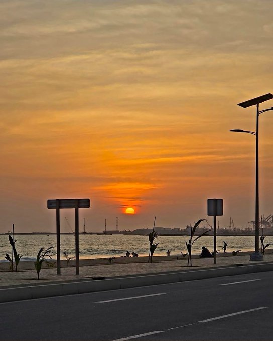 A sunset scene over a coastal road with the orange sun setting over the water, silhouetted street lamps, distant port cranes, and people on the shore.