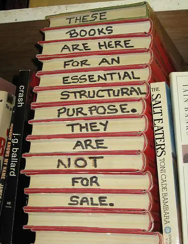 A stack of books with red and cream spines displays handwritten text reading 'THESE BOOKS ARE HERE FOR AN ESSENTIAL STRUCTURAL PURPOSE. THEY ARE NOT FOR SALE.'