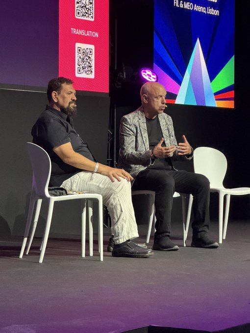 Two men sit on white chairs on a stage during an event; one wears a black shirt and the other a patterned silver blazer, with a screen behind them displaying 'FTL & MEO Arena, Lisbon' and a 'TRANSLATION' QR code.