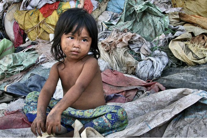 A young child with dirt on their face sits among large piles of worn sacks, cloth, and refuse, looking directly toward the camera.