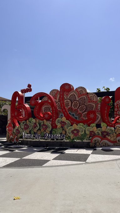 An outdoor photo of a colorful red floral-themed display spelling out a word with the text 'IMAGINE THE PARADISE' visible at the bottom, set against a clear blue sky with mountains in the background.