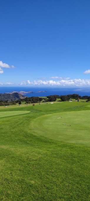 A scenic view of a coastal golf course with a green in the foreground, rolling hills, the ocean, and islands visible under a blue sky with scattered clouds.