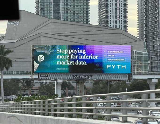 A digital billboard displays an advertisement for PYTH with the text 'Stop paying more for inferior market data' listing features like 'EVERY ASSET CLASS', 'ONE API', and 'ZERO LICENSING FEES', located above a highway in front of the Adrienne Arsht Center.