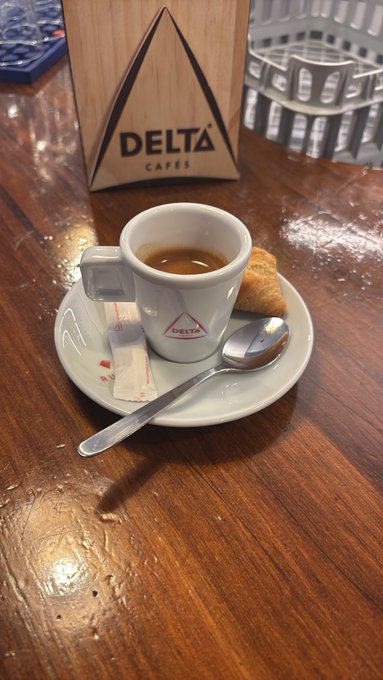 A small espresso cup with the Delta Cafes logo sits on a white saucer with a spoon, accompanied by sugar packets and a small biscuit on a wooden table.