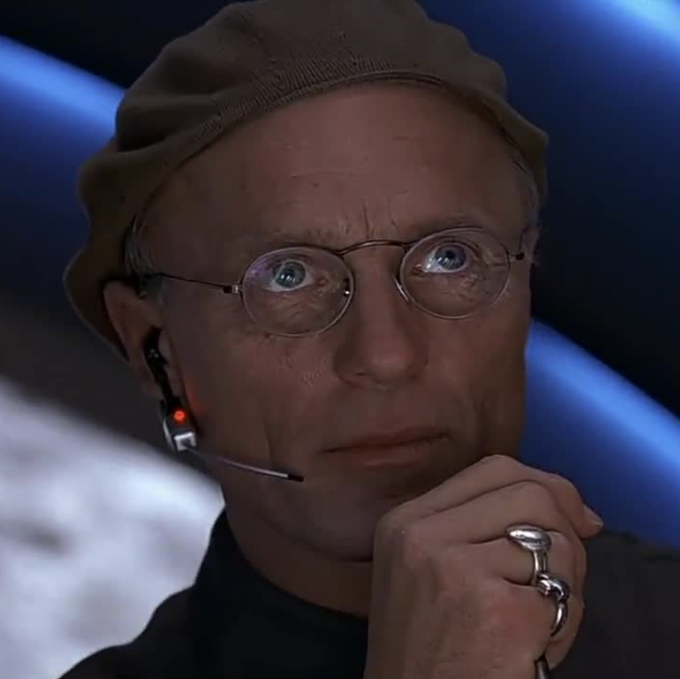A bald man with round wire-frame glasses, a brown beanie, and a headset microphone stares thoughtfully with wide eyes, his hand resting near his chin while wearing a silver knuckle ring. His skeptical, calculating expression perfectly mirrors the manipulative tone of the caption, which suggests orchestrating Bitcoin's rise to $80,000 before crashing it to align with the four-year cycle theory.