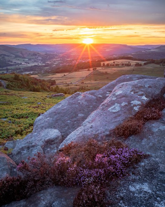 A scenic landscape photograph showing a vibrant sunset over a hilly, green countryside with large rocks and purple heather in the foreground.