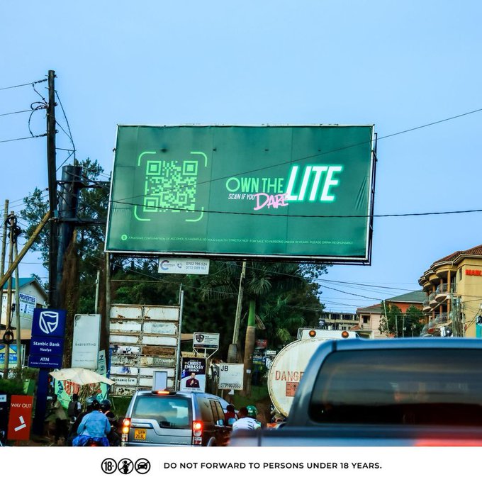 The image shows a busy street scene with cars driving by and people walking along the sidewalk. There are two billboards prominently displayed in the background - one green billboard that says "OWN THE LITE" and another white billboard that has an image of a person on it, possibly advertising something related to the green billboard.

In addition to the billboards, there is also a car driving down the street with its rear view mirror visible. The scene captures the hustle and bustle of city life, with various elements such as traffic lights, buildings, and people going about their day-to-day activities.