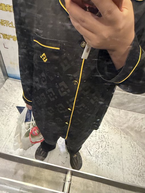A mirror selfie showing a person wearing a black patterned jacket or coat with yellow piping, holding shopping bags in what appears to be a store with tiled flooring.