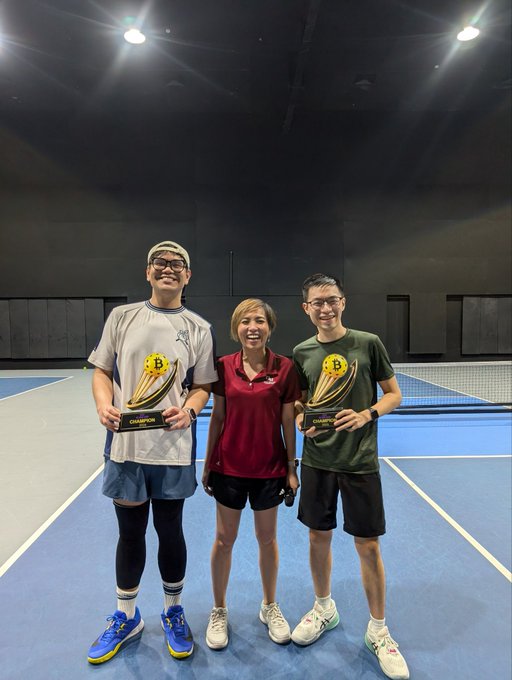 Three people posing on an indoor tennis/pickleball court, with two men on either side holding 'CHAMPION' trophies and all three smiling at the camera.