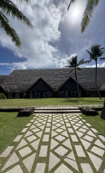 A tropical resort building with a thatched roof is framed by palm trees, approached by a geometric stone walkway across a grassy lawn under a partly cloudy sky.