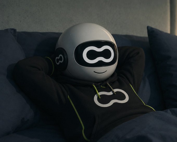 A figure wearing a black hoodie with a yellow-green infinity logo lounges on a dark couch, wearing a white spherical helmet featuring a digital infinity-style visor and a smile.